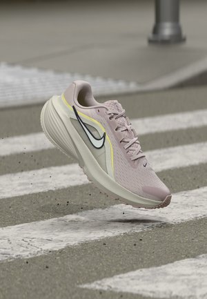 Chaussure de course Nike rose clair avec semelle blanche et swoosh noir, suspendue au-dessus d'un passage piéton sur une route asphaltée.