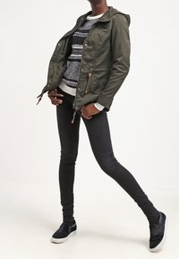 Chaqueta verde oliva con capucha y dos bolsillos laterales, superpuesta a un suéter estampado. Jeans skinny negros y zapatos slip-on azul marino completan el look.