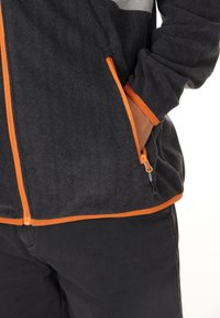 Persona che indossa una giacca nera con zip, decorata con bordini e zip arancioni, con una mano nella tasca zip, abbinata a pantaloni neri.
