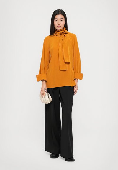 Blusa rustica arancione con dettaglio al collo a fiocco, abbinata a pantaloni neri a gamba larga. Il modello tiene una pochette color crema.