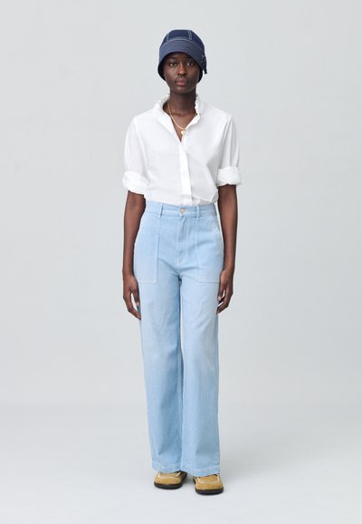 Chemise blanche à boutons avec manches retroussées, pantalon large rayé bleu clair, chapeau seau navy à rayures et chaussures beiges.