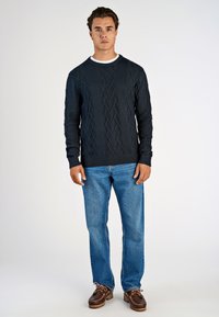 Marineblauer Kabelstrickpullover mit texturiertem Muster, Rundhalsausschnitt, gerippten Bündchen, kombiniert mit hellblauen Jeans und braunen Mokassin-Schuhen.