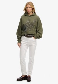 Sudadera extragrande de color verde oliva con diseño de alas y lentejuelas, combinada con jeans blancos y mocasines marrones. Textura suave, ajuste casual.