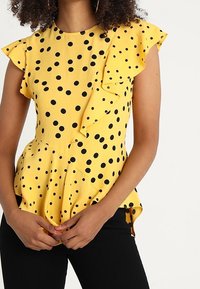 Žlutý top s černými puntíky, volánkovými rukávy a peplum střihem. Obsahuje knoflíkové detaily na boku a strukturovanou tkaninu.