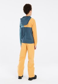 Felpa con cappuccio in tonalità turchese e gialla, con un cappuccio e vari blocchi di colore. Abbinata a pantaloni larghi e brillanti gialli e scarpe nere.