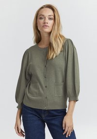 Cardigan verde, con texture, colletto rotondo, chiusura con bottoni, maniche a sbuffo e due tasche frontali, abbinato a jeans scuri.