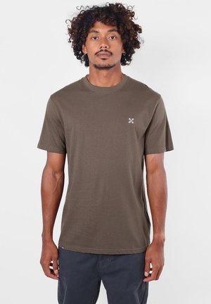 Homme aux cheveux bouclés portant un t-shirt marron uni à manches courtes et un pantalon foncé, debout devant un fond uni clair.
