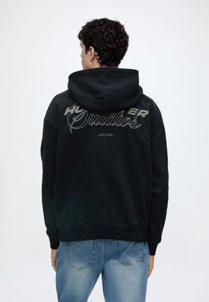 Jeune homme aux cheveux bouclés portant un sweat à capuche noir avec le texte "Holster Studios" dans le dos et un jean bleu clair, debout, de dos.
