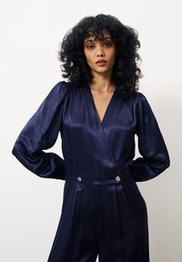 Combinaison en satin navy avec de longues manches bouffantes, décolleté croisé, et boutons décoratifs en argent à la taille. Texture lisse, coupe cintrée.