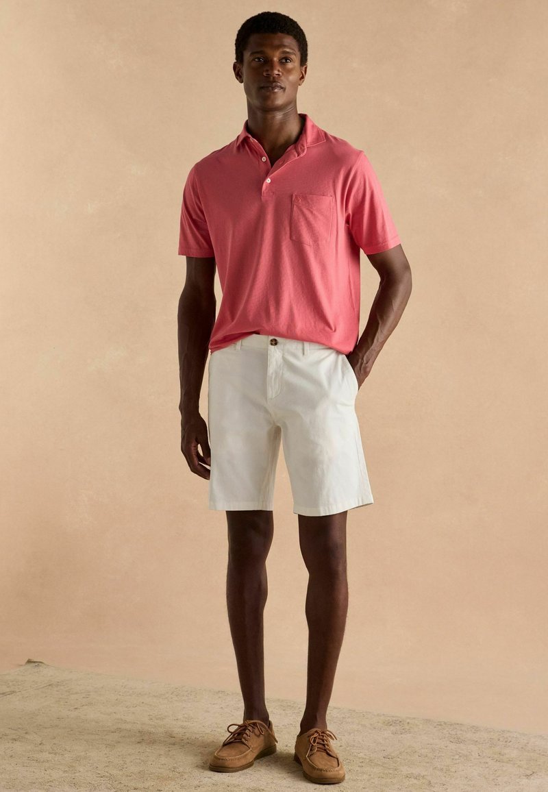 Lühikeste varrukatega roosa polo, millel on tasku, koos valgete lühikesed pükstega. Mudel kannab pruune nööbisaapaid. Neutraalne taust toob riietuse esile.