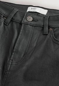 Jean skinny noir avec taille mi-haute, présentant des coutures contrastées, une fermeture à bouton sur le devant et une étiquette en tissu avec des instructions d'entretien.
