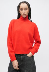 Czerwony sweter z golfem w miękkim dzianinowym materiale, krótki fason z prążkowanymi mankietami i ściągaczem, połączony z czarnymi spodniami z wysokim stanem.