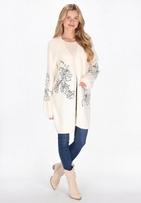 Gehaakte cardigan in crème, met bloemendessins in blauw en rood op de mouwen. Gepaard met donkere skinny jeans en beige enkellaarzen.
