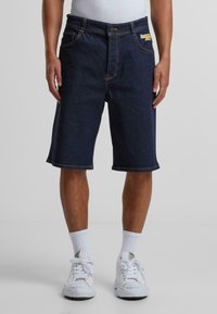 Shorts en denim bleu foncé avec logo brodé jaune, longueur mi-cuisse, cinq poches et couture contrastée, associés à des baskets blanches.