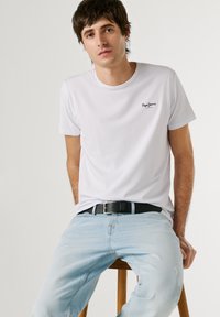 Fiatal férfi ül egy fa széken, fehér Pepe Jeans pólót és világoskék farmert visel, kezei a csípője mellett pihennek.