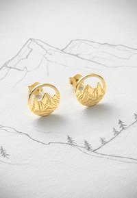 Guldcirkulära örhängen med en bergslandskapsdesign som har graverade träd och berg, mot en ljus strukturerad bakgrund.
