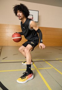 Giocatore di basket in canotta nera senza maniche e pantaloni corti, che dribbla un pallone da basket rosso. Indossa sneakers multicolori con accenti gialli e rossi.