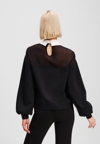 Sweatshirt noir avec un dos en tissu transparent, col blanc, découpe en forme de trou de clé au niveau du cou et manches oversized. Texture douce et coupe décontractée.