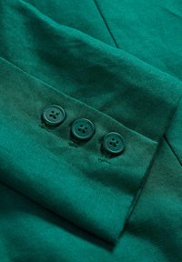 Tessuto verde con una superficie testurizzata, che mostra tre bottoni scuri su un polsino abbottonato. I dettagli delle cuciture sono visibili. Nessun motivo o accento.