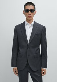 Karbonfarbener Anzug mit strukturierten Schultern, Revers und zwei vorderen Taschen, kombiniert mit einem weißen Hemd und schwarzer Sonnenbrille.