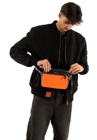 Chaqueta bomber negra sobre una camiseta gris, sosteniendo un bolso bandolera naranja y negro con cremallera y cadena como correa. Tela texturizada.