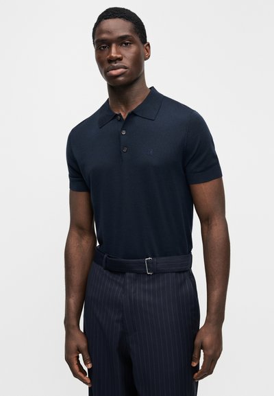 Μπλε ναυτικό πόλο με κοντά μανίκια, κλασικό γιακά και τρία κουμπιά στο εμπρός μέρος, συνδυασμένο με μπλε ναυτικό παντελόνι με ρίγες και ζώνη στο ίδιο χρώμα.