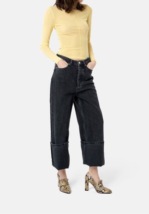 Kvinde iført korte sorte viddebenede jeans, gul langærmet top og højhælede sko med slangeskindsprint, hænderne i lommerne.