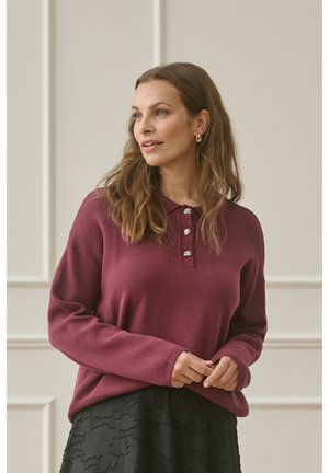 Pull en maille bordeaux à manches longues, col boutonné, et texture douce, associé à une jupe noire à motifs.