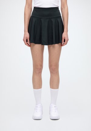 Une personne porte une jupe de tennis plissée noire avec un haut blanc, des chaussettes montantes et des chaussures de sport blanches.