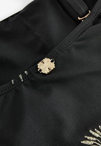 Čierna látka s detailným šitím, obsahujúca malú ozdobnú zlatú sponu a vyšívaný zlatý kvetinový vzor.