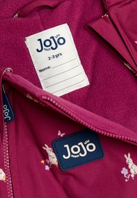 Chaqueta de forro polar en rosa profundo con patrones de flores y conejos; presenta una etiqueta de marca azul y una etiqueta de cuidado de la tela blanca con un espacio para el nombre.
