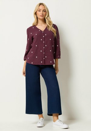 Vrouw draagt een bordeauxrode blouse met knopen en kleine witte bloemenprint, donkere wijde spijkerbroek en witte sneakers, staand tegen een effen achtergrond.