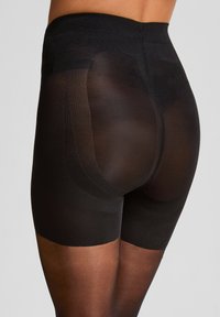 Frau in schwarzen, durchsichtigen Formshorts, die die Konturen des Gesäßes und die Oberschenkelbedeckung betonen, vor einem hellen Hintergrund.