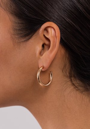 Gros plan sur l'oreille d'une femme aux cheveux foncés portant une épaisse boucle d'oreille en or brillant.