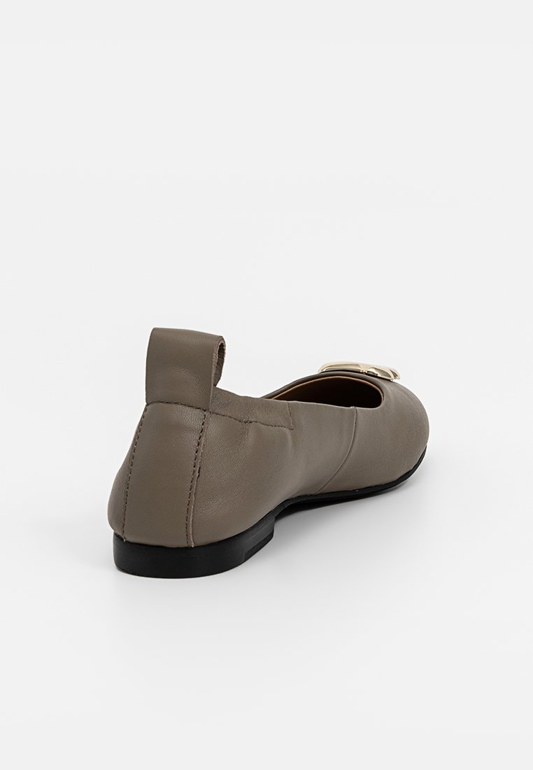 Płaskie buty skórzane w kolorze taupe z czarną podeszwą, z tylnym uchwytem i złotym zapięciem z przodu, ustawione w kącie, aby pokazać tylny i boczny widok.