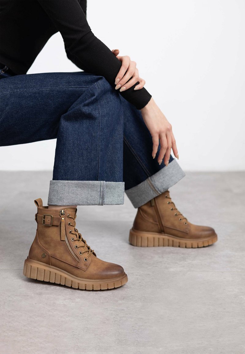 Botas de cuero marrón hasta el tobillo con suela de goma gruesa, diseño con cordones, cremallera lateral y detalle de correa ajustable. Pantalones de mezclilla con dobladillos enrollados.