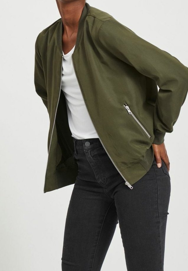 Veste bomber vert olive en tissu lisse et léger. Dotée d'une fermeture éclair à l'avant et de poches latérales. Portée sur un haut blanc et un jean noir.