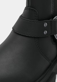 Botín negro de tobillo con material de cuero suave, correa ajustable con acentos metálicos y costuras detalladas a lo largo de las uniones.