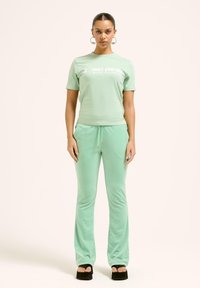 Mėtos žalios spalvos "Juicy Couture" marškinėliai ir atitinkamos sportinės kelnes iš minkštos medžiagos. Marškiniai pasižymi balto logotipo tekstu; kelnės turi elastingą juosmenį ir raištelius.