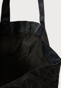 Čierna nylonová taška cez rameno s hladkou textúrou, jemnou potlačou loga a kontrastnými lesklými remienkami. Interiér má jednofarebný dizajn.