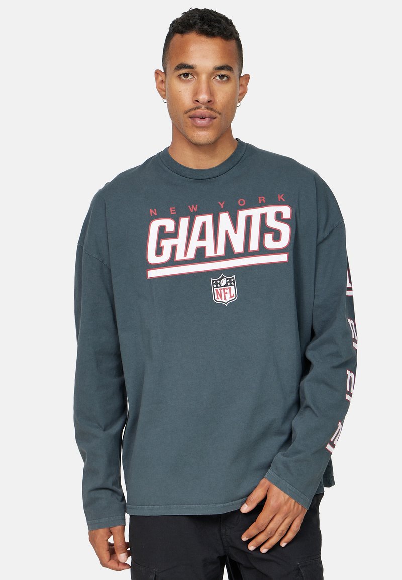 Pitkähihainen crewneck-t-paita tummansinisellä pohjalla, jossa on "New York Giants" -grafiikkaa valkoisena ja punaisena edessä ja hihassa; puuvillamateriaali.