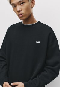 Sort oversized sweatshirt med ribbede krave og manchetter, prydet med et lille hvidt logo på brystet. Blødt stof, afslappet pasform.
