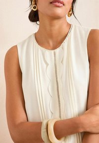 Sleeveless ivory blouse with scalloped neckline, vertical pleats, and button detail. Features a smooth texture and elegant design accents.