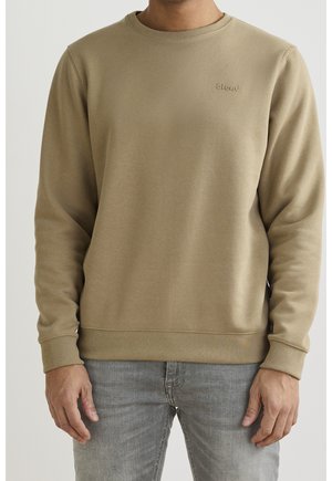 Homme portant un sweat-shirt uni marron clair à col rond avec poignets et ourlet côtelés, associé à un jean gris clair.