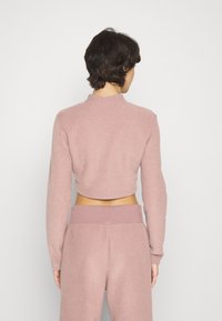 Avkortad långärmad topp i mjuk rosa tyg, med hög krage och avspänt snitt, kombinerad med matchande byxor som har ett enkelt midjeband.