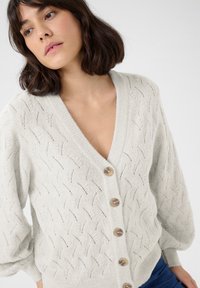 Cardigan tricoté gris clair avec un col en V, présentant un motif texturé et de grands boutons beiges sur le devant. Tissu doux, coupe ample.