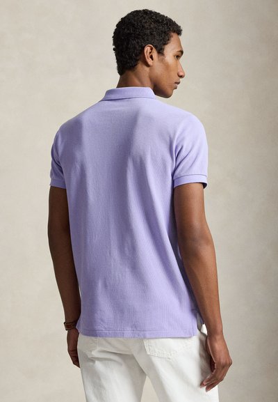 Hombre con pelo rizado que lleva una camiseta polo de manga corta lavanda y pantalones blancos, de espaldas sobre un fondo liso.