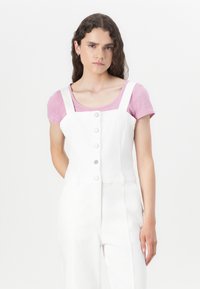 Mono de mezclilla blanco con escote cuadrado, cierre frontal de cinco botones y tirantes ajustables. Superpuesto a una camiseta de rayas rosa claro.