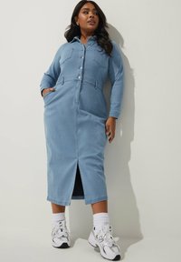 Ljusblå denimklänning med långa ärmar, knappstängning, bröstfickor och en slits till låret. Kombinerad med vita sneakers och strumpor.