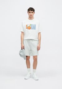 Άσπρο γραφικό t-shirt με την επιγραφή «AIR JORDAN», γκρι σορτς και γκρι καπέλο. Το σύνολο ολοκληρώνουν λευκά αθλητικά παπούτσια και χαμηλές κάλτσες.